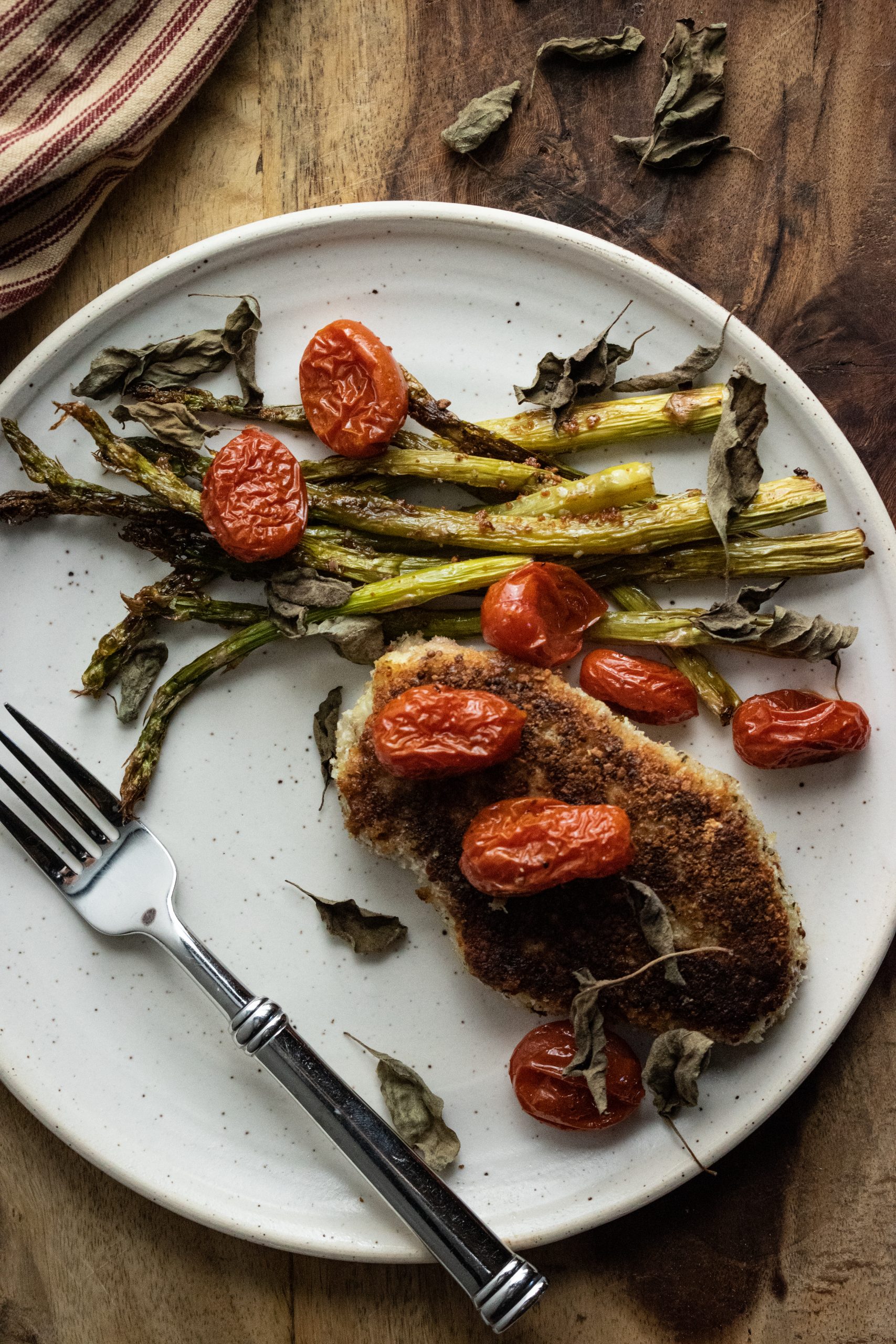 crispy panko coated porkchops with roasted asparagus and burst cherry tomatoes on a white plate on a wooden cutting board