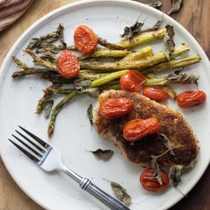 easy pork chops encrusted in a panko mix on a white plate with roasted asparagus and cherry burst tomatoes