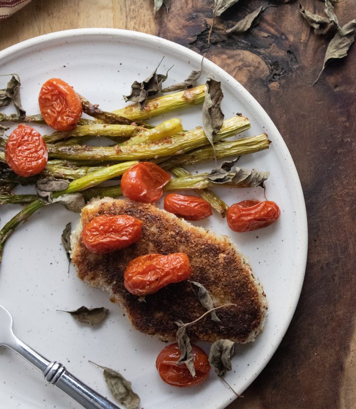 birds eye view of crispy porkchops on a white plate with asparagus and roasted cherry tomatoes