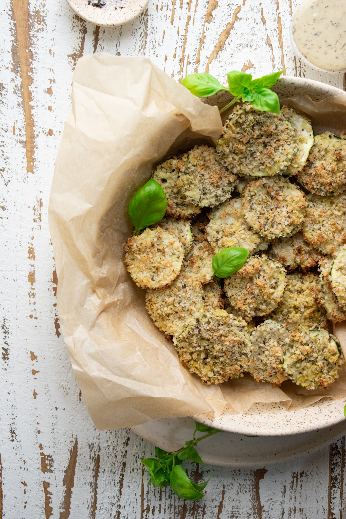 close up of oven baked zucchini chips in a bowl filled with parchment paper on a wooden white board