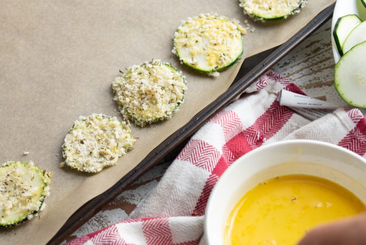 zucchini chips covered in panko breadcrumbs lined up on a baking tray