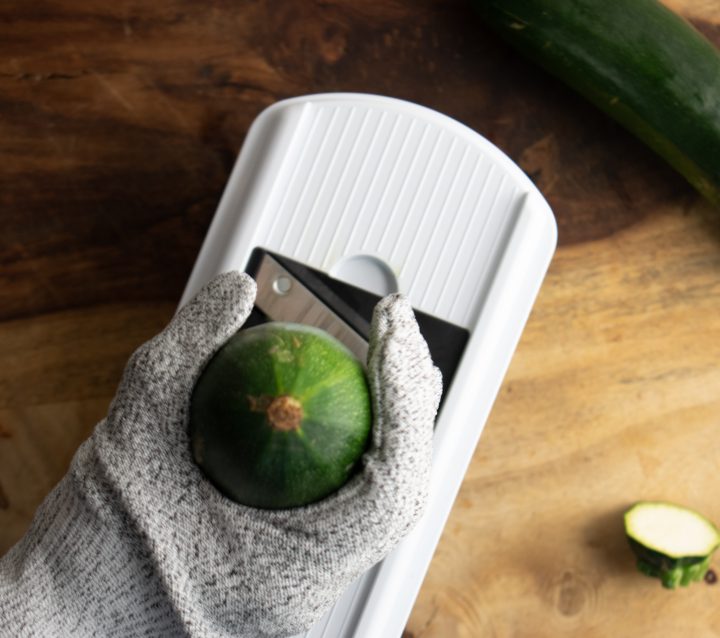 mandoline slicer with a hand wearing a safety glove and holding a zucchini to slice