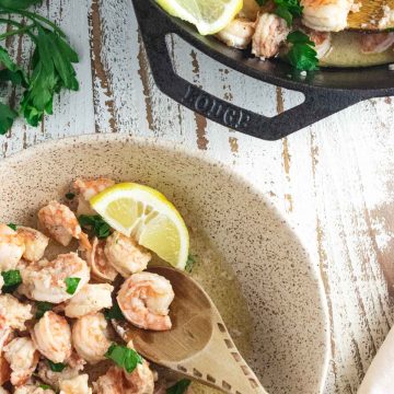 Shrimp scampi in a cream colored bowl with some fresh parsley scatterd on top and a wooden slotted spoon on the side and a lemon wedge placed inside.