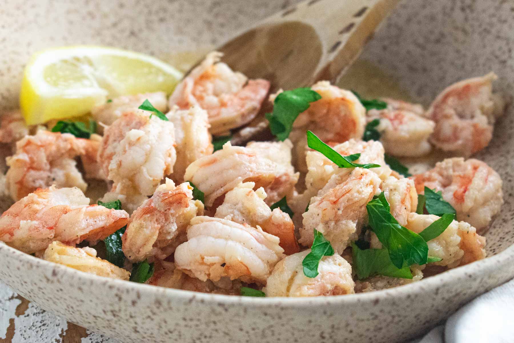 a close up of shrimp scampi in a cream colored bowl topped with fresh parsley and a wooden spoon slotted in the side and a fresh lemon slice