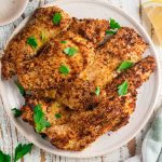 A birds eye view of crispy chicken cutlets on a cream colored plate with fresh parsley scattered on top and a slice of lemon on the side.