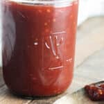 BBQ sauce in a glass jar on a wooden table with a spoon laid next to it with a dab of bbq sauce on it.