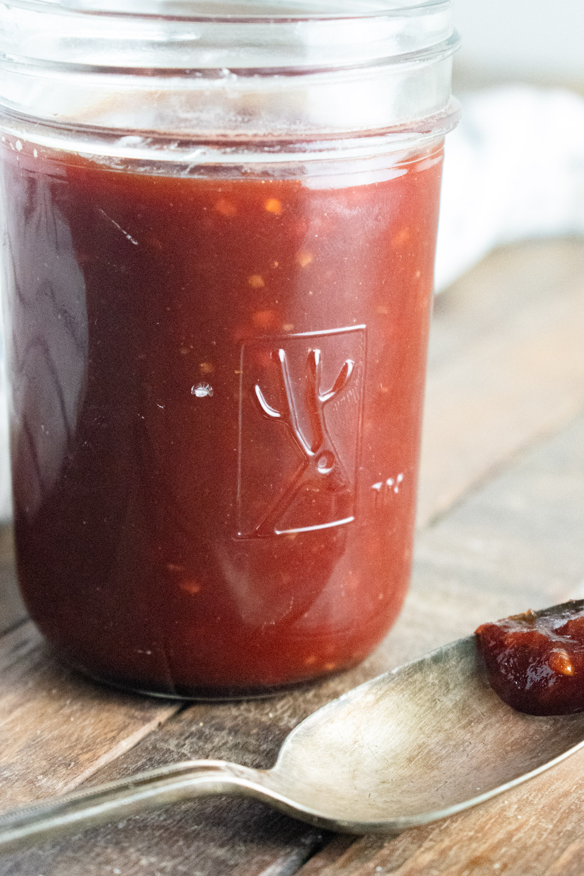 BBQ sauce in a glass jar on a wooden table with a spoon laid next to it with a dab of bbq sauce on it.
