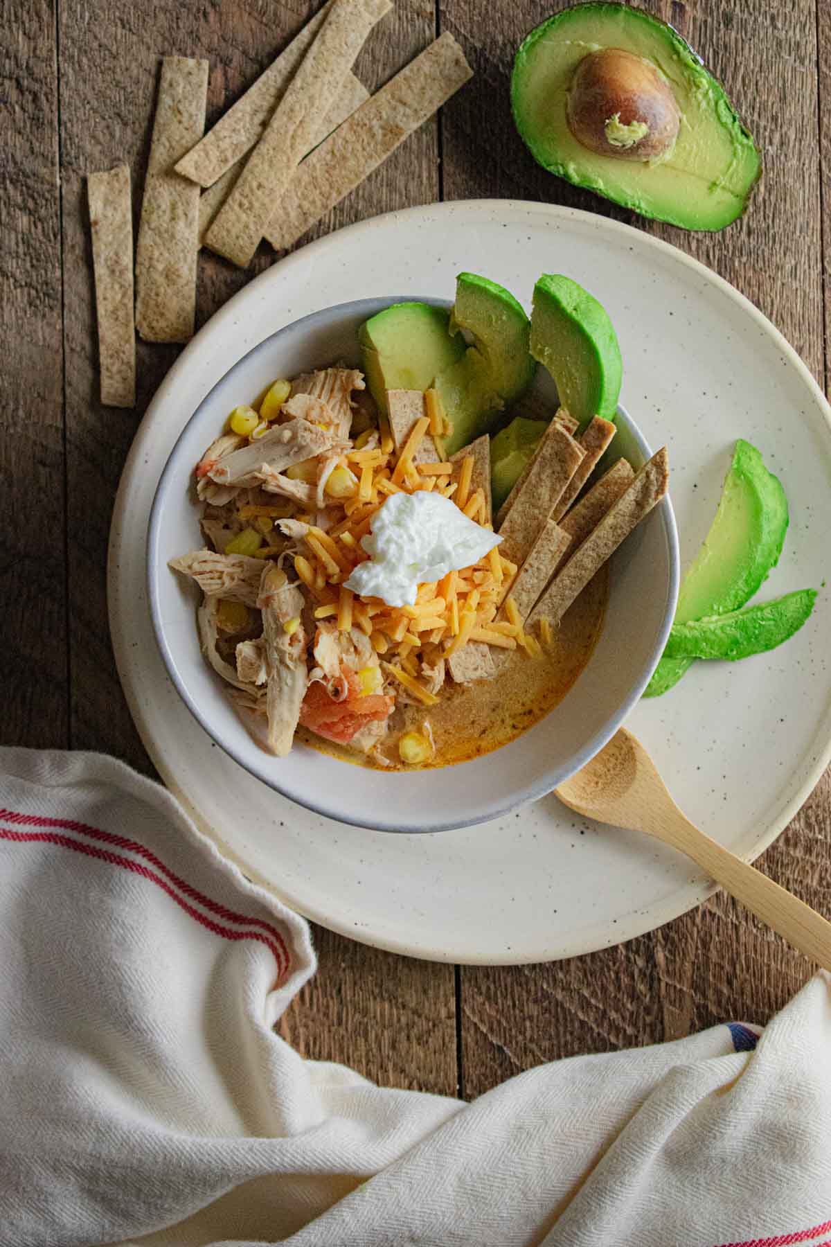 slow cooker healthy chicken tortilla soup in a white bowl on a wooden table with tortilla strips and an avocado next to the bowl