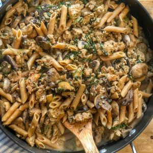 chicken sausage with mushroom penne in a large frying pan with a wooden spoon