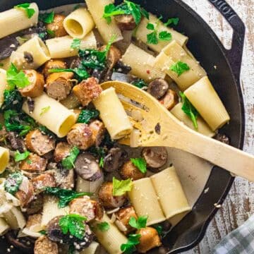 Chicken sausage and pasta in a cast iron skillet with garlic, onion, and baby spinach in a creamy sauce with a wooden slotted spoon on the side.