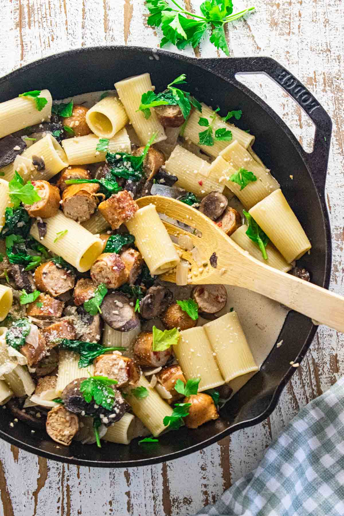 Chicken sausage and pasta in a cast iron skillet with garlic, onion, and baby spinach in a creamy sauce with a wooden slotted spoon on the side. 