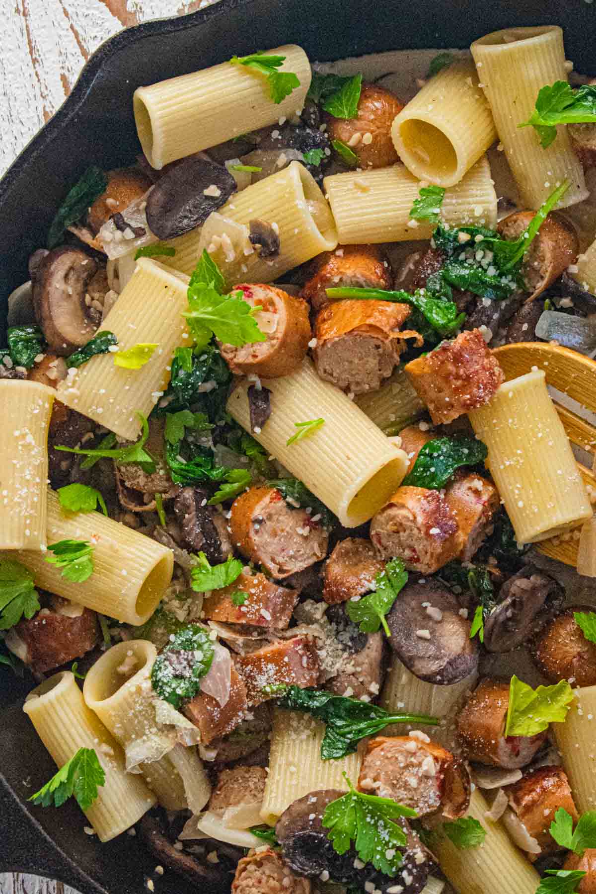 A close up of chicken sausage and pasta in a cast iron skillet with garlic, onion,and baby spinach.