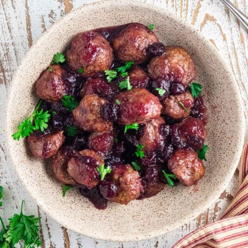 A birds-eye view of juicy cranberry meatballs in a cream colored mixing bowl on a white wooden board with a brown and white striped napkin next to the bowl.