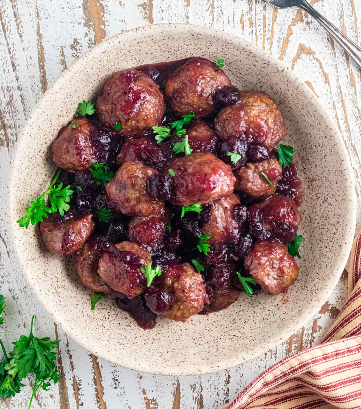 A birds-eye view of juicy cranberry meatballs in a cream colored mixing bowl on a white wooden board with a brown and white striped napkin next to the bowl.