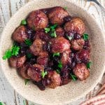 A birds eye view of juicy cranberry turkey meatballs in a cream colored bowl on a white wooden table with a brown and white striped napkin next to the bowl.