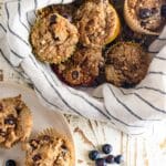 Healthy Blueberry muffins in a basket with a striped napkin laid inside and a few muffins next to the basket on a plate.
