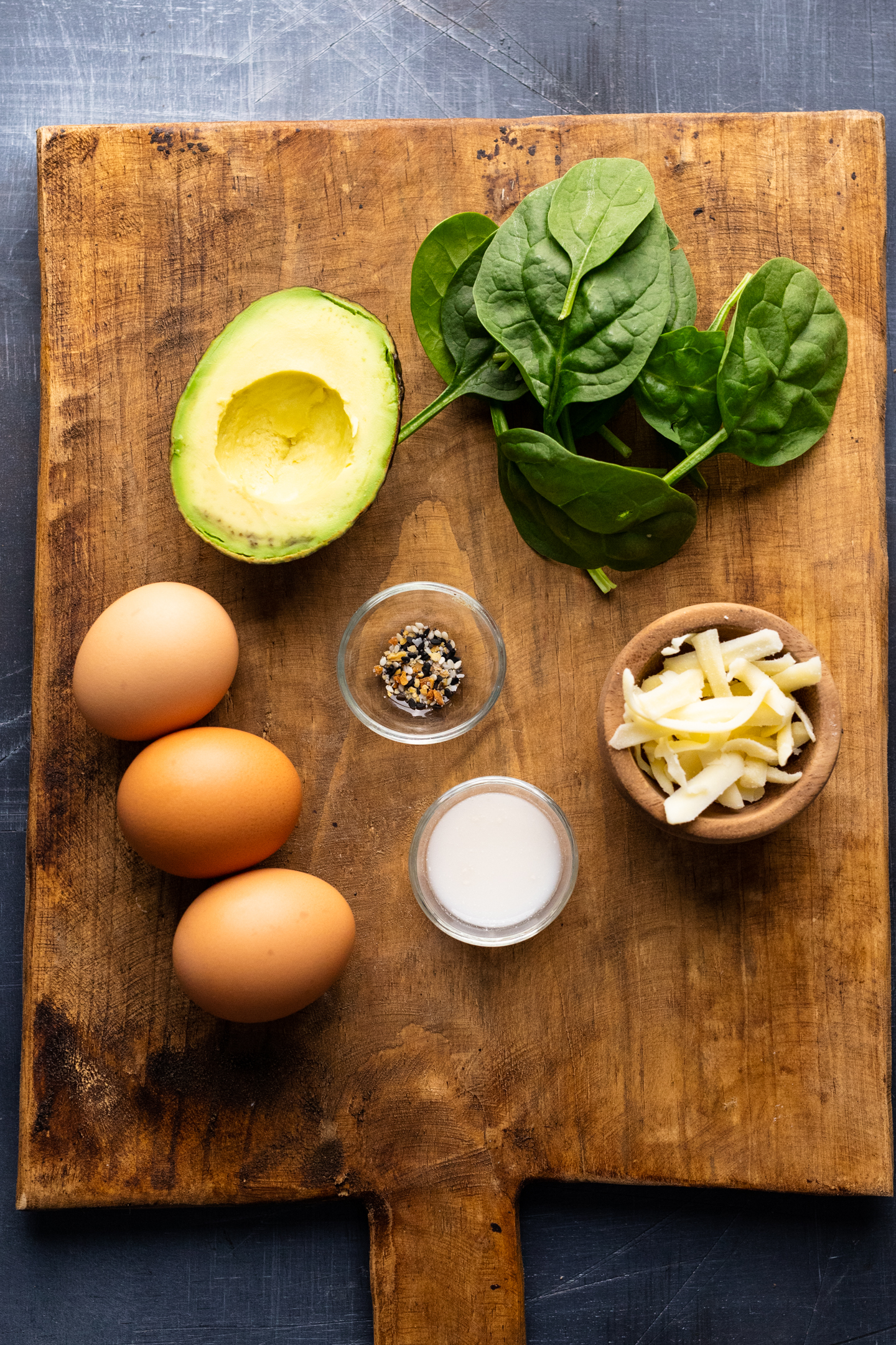 Ingredients for a spinach and avocado omelette arranged on a wooden board that's sitting on a gray background.