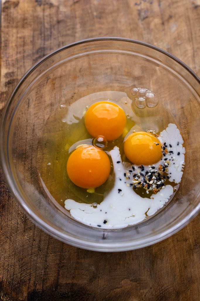 A clear glass bowl with eggs, milk, and seasoning. The bowl is arranged on a wooden board.