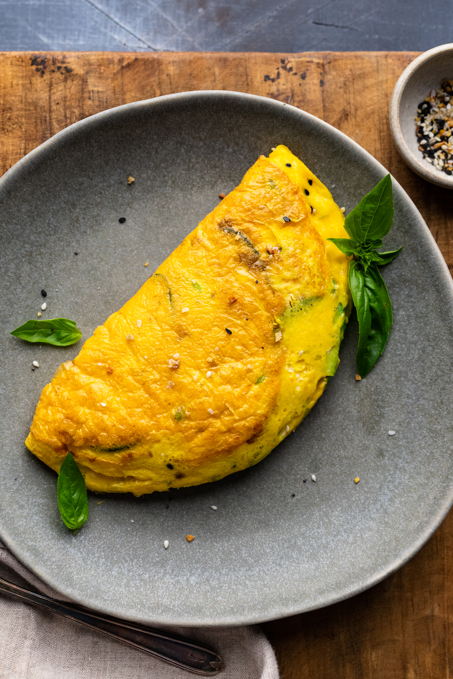 A gray plate with an avocado omelette on it with fresh herbs arranged around it. The plate is sitting on a wooden board.