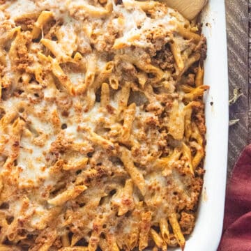 Baked ziti in a white casserole dish on a wooden table with a wooden spoon slotted into the side.