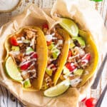 Mahi Mahi fish tacos in a cream colored dish with 3 limes on top and cherry tomatoes scattered to the side.