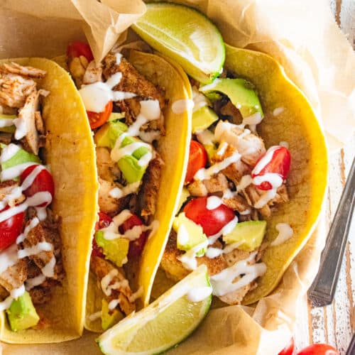 Close up of 3 fish tacos with limes and cherry tomatoes scattered about.