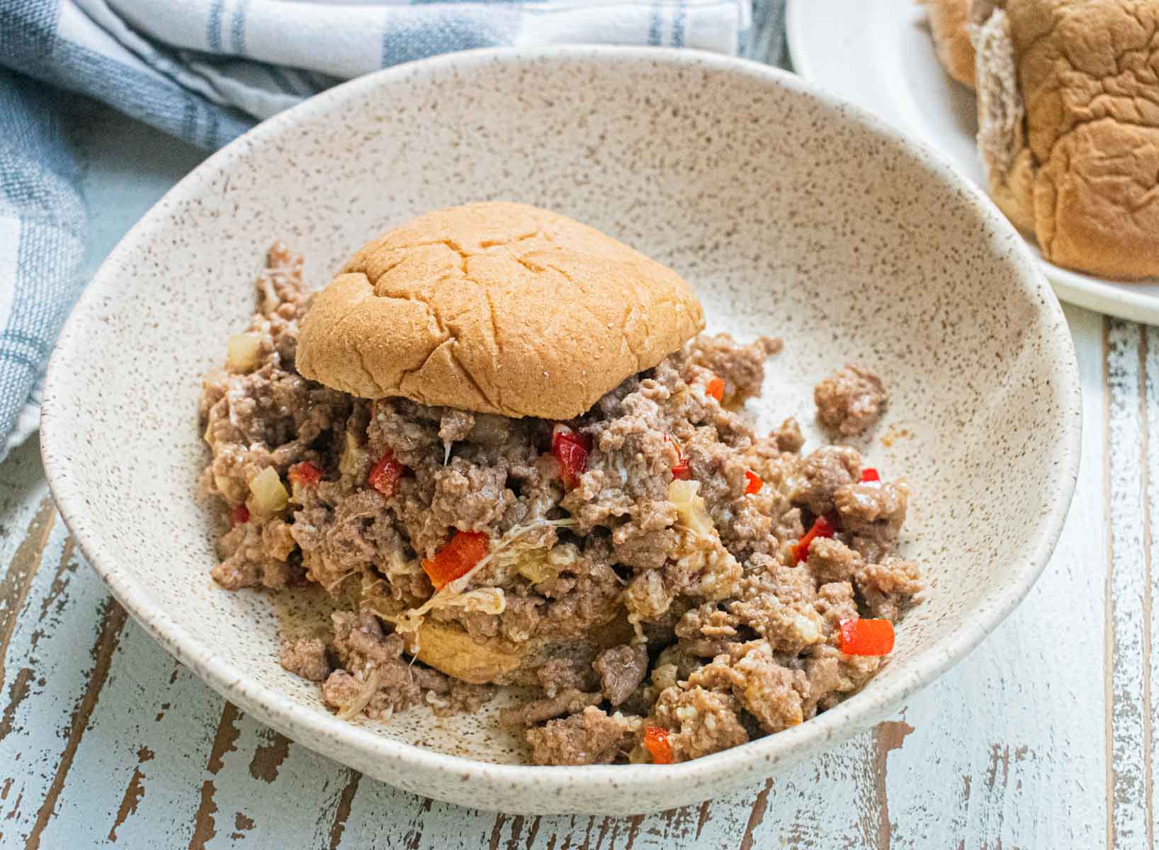 A close up of ground turkey sloppy joes in a white bowl on a white wooden table with a checkered napkin in the background.