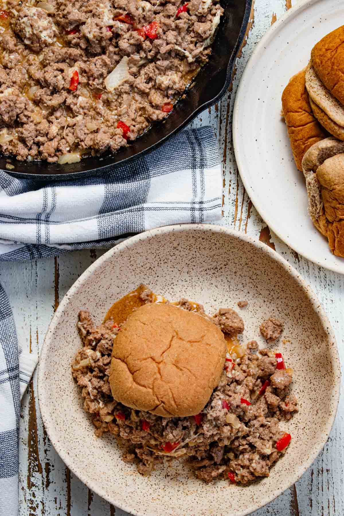 Sloppy joes assembled on a whole wheat bun on a white plate with a cast iron skillet of meat sitting next to a plate of buns.