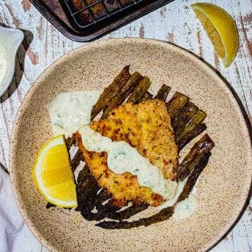 Birds-eye view of salmon on a cream colored plate with tartar sauce drizzled on top and a slice of lemon on the side.