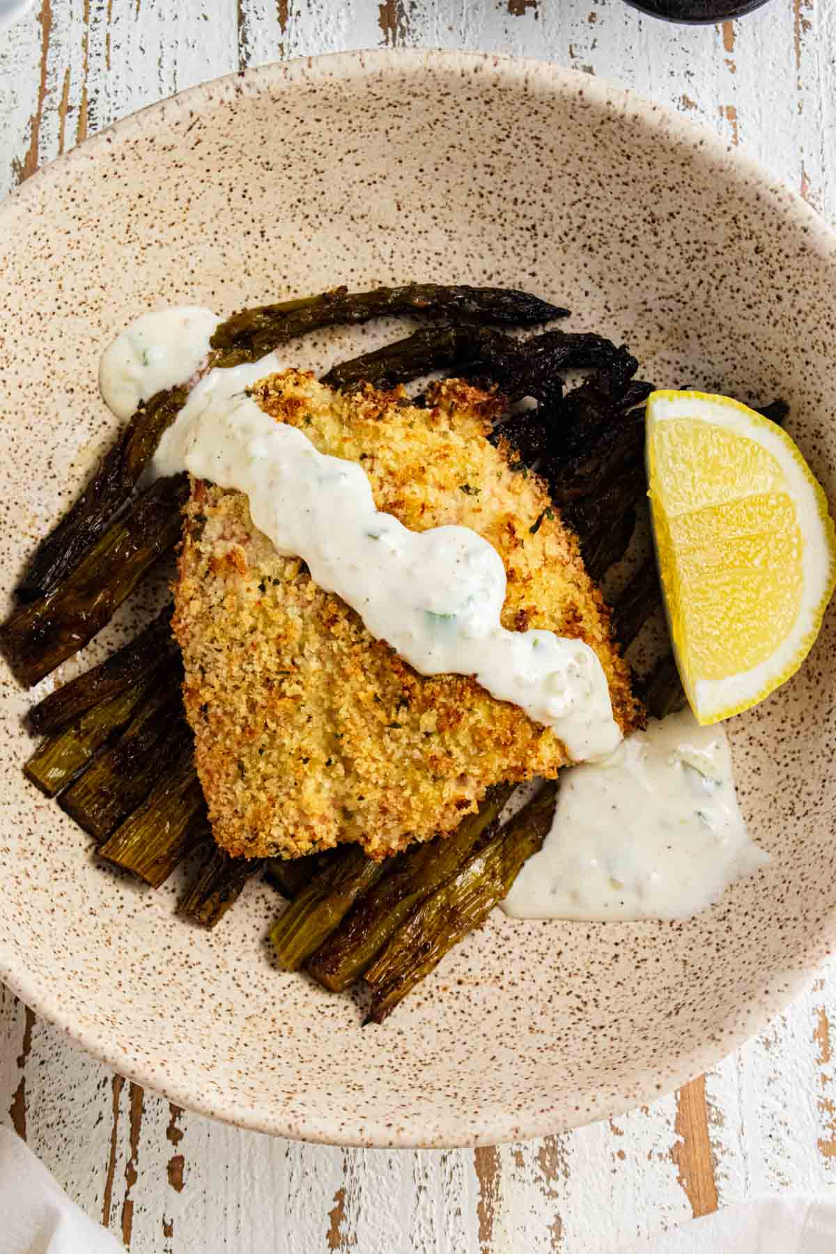 A close up of panko crusted salmon on a cream colored plate with tartar sauce drizzled on top and a slice of lemon on the side.