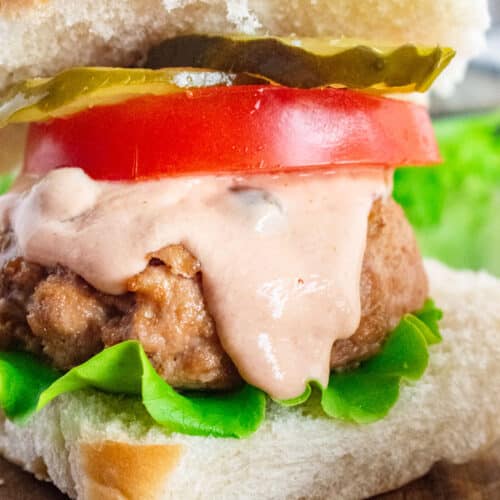 A close up of a turkey burger on a whole wheat bun with lettuce, tomato, pickle, and burger sauce dripping down.