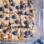 Blueberry Oat Peanut Butter Bars in a clear dish on a wooden table with blueberries scattered about.