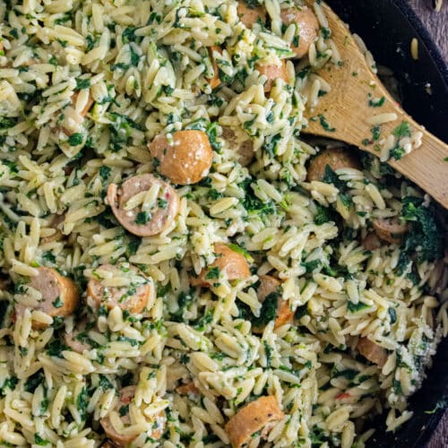 Close up of chicken sausage orzo in a cast iron skillet with a wooden spoon slated in the dish.
