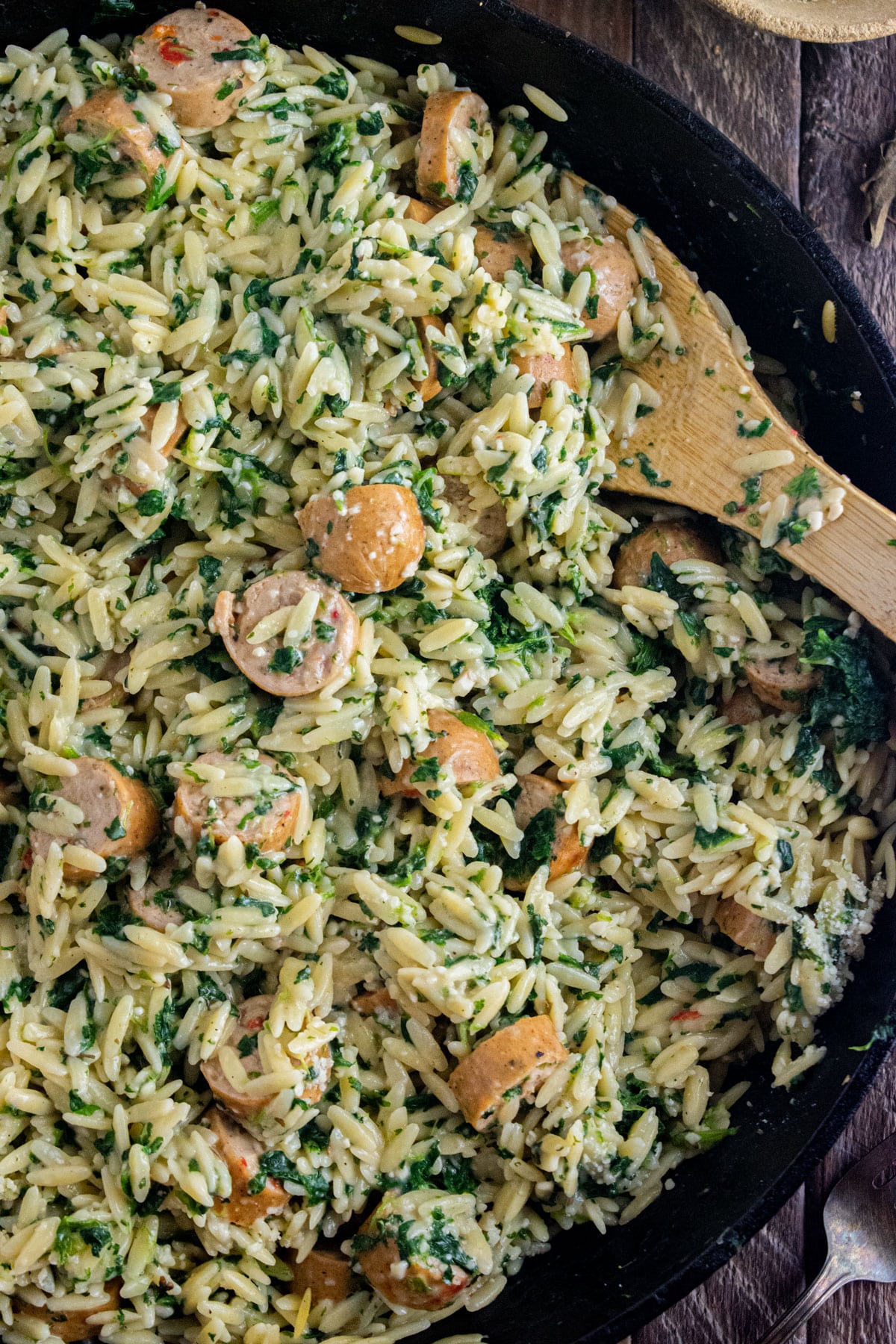 Close up of chicken sausage orzo in a cast iron skillet with a wooden spoon slated in the dish.