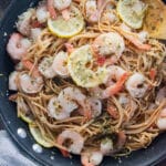 Shrimp and linguini in a pan with a wooden spoon slotted in the side and lemon slices scattered on top.