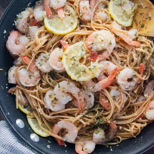 Shrimp and linguini in a pan with a wooden spoon slotted in the side and lemon slices scattered on top.