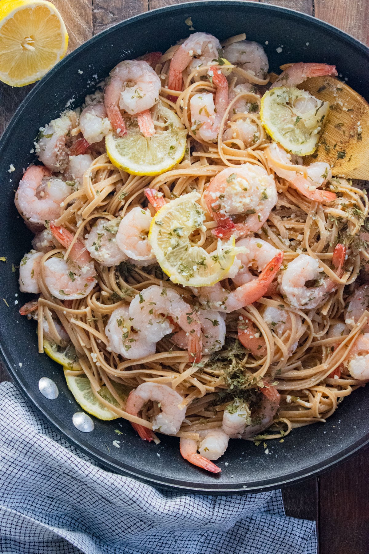 Shrimp and linguini in a pan with a wooden spoon slotted in the side and lemon slices scattered on top.