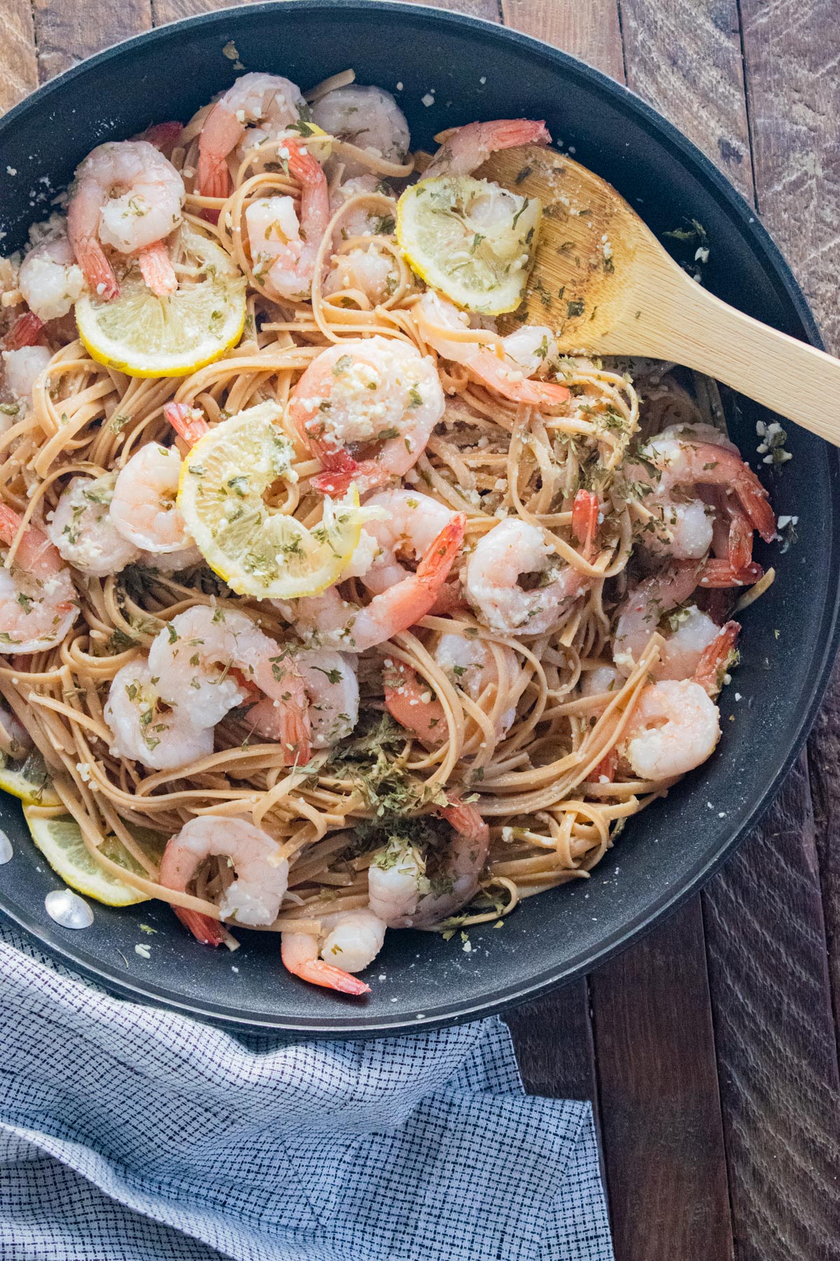 Shrimp and linguini in a skillet with a wooden spoon slotted inside and lemon slices scattered on top.