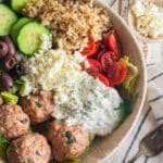 A close up of Greek chicken meatballs in a dish with tomatoes, cucumbers, olives, feta cheese, rice, and Tzatziki sauce.