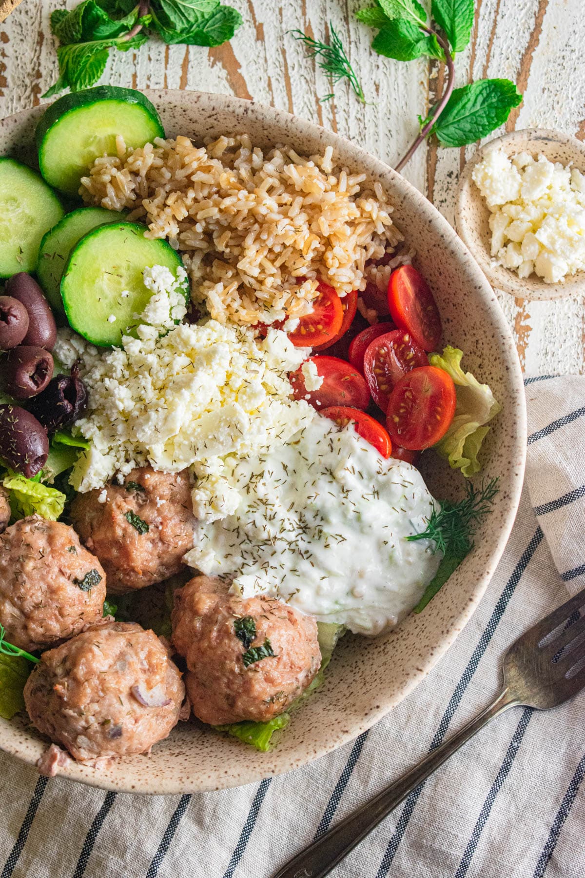 A close up of Greek chicken meatballs in a dish with tomatoes, cucumbers, olives, feta cheese, rice, and Tzatziki sauce.