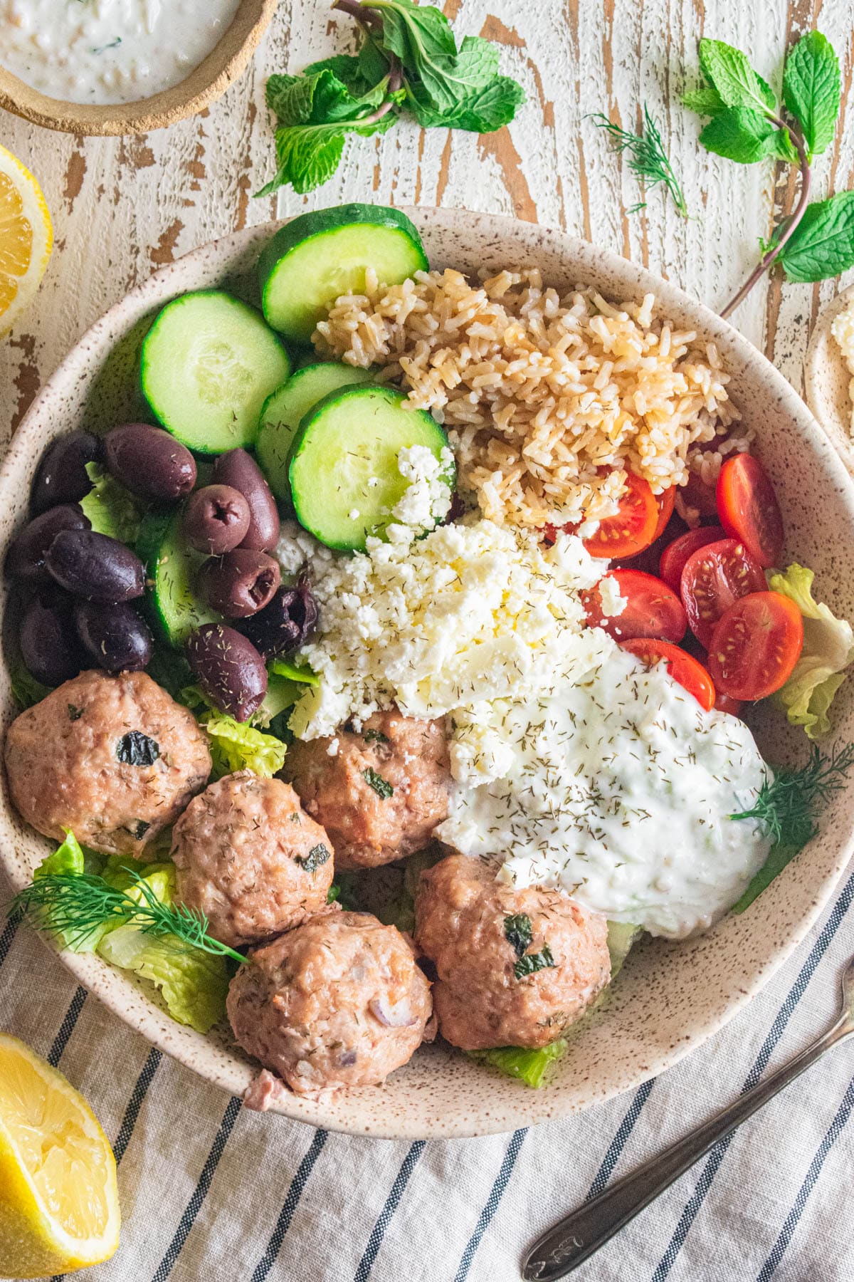 Greek chicken meatballs in a bowl with cucumbers, olives, tomatoes, rice, feta cheese, and Tzatziki sauce.