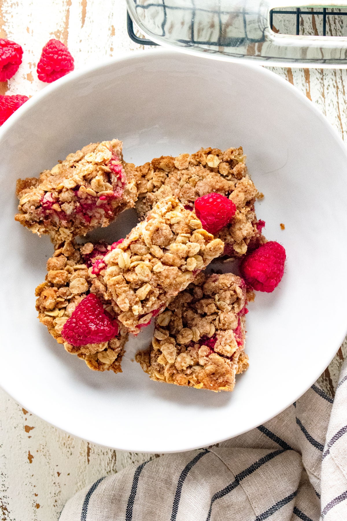 Berry oatmeal bars in a white bowl on a wooden table with a blue and white striped napkin laid next to the dish and raspberries scattered on top.