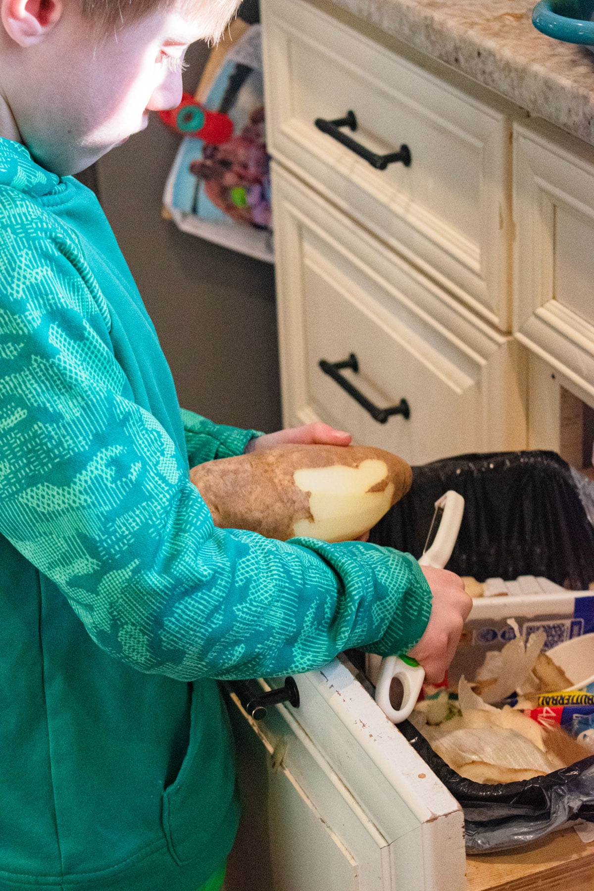 A little boy using a kid-friendly peeler and peeling a potato into the garbage.
