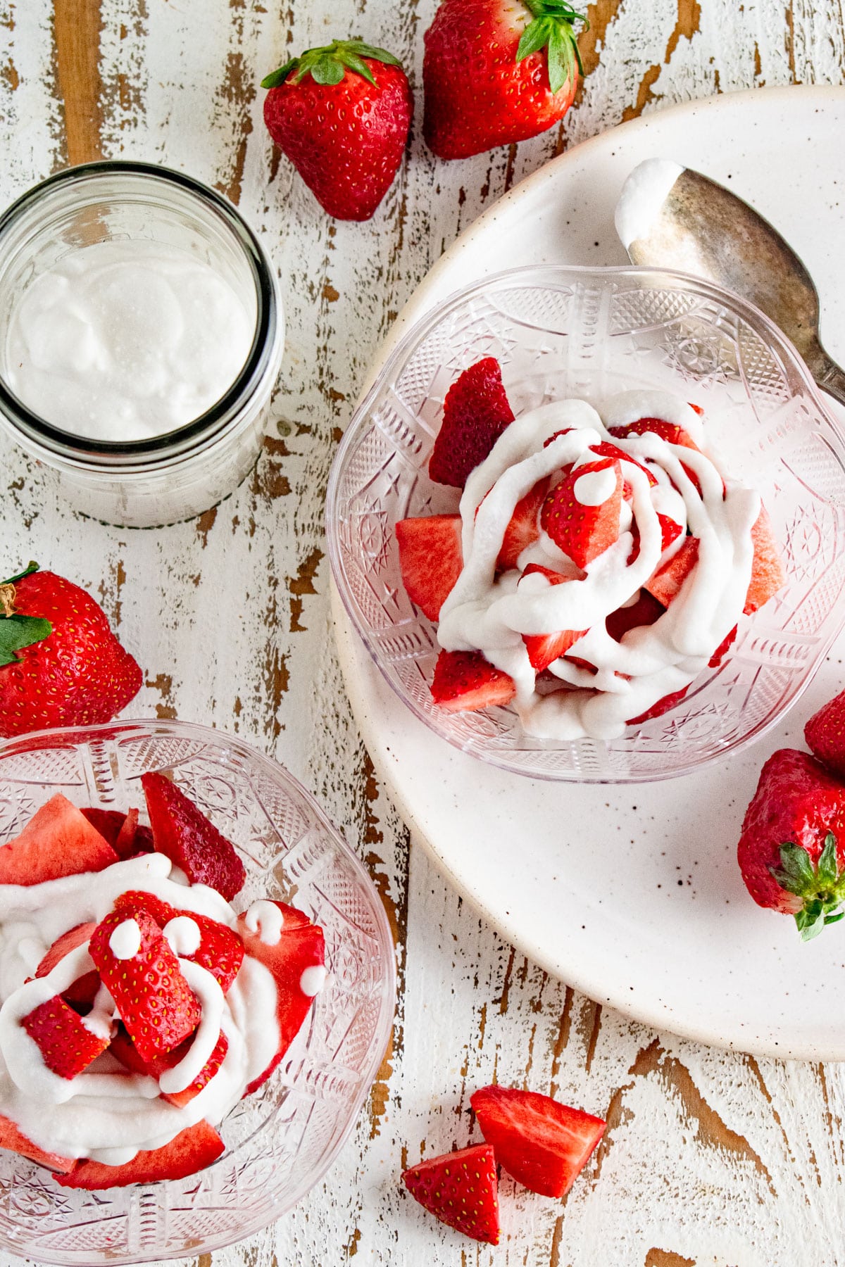 Two clear dishes on a white wooden table with strawberries and whipped cream on display.