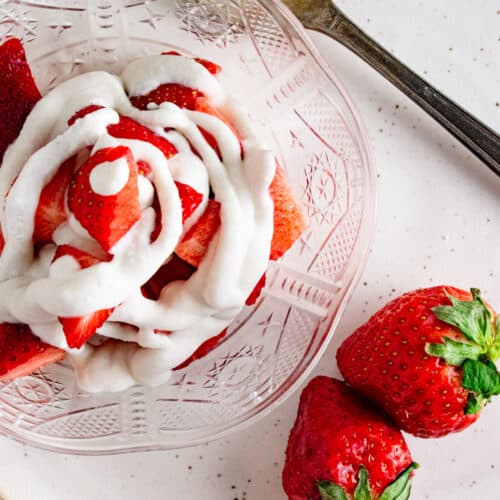 A close up of coconut whipped cream in a clear dish with strawberries laid next to the dish.