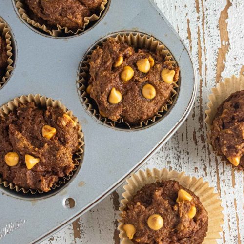 A close-up of pumpkin butterscotch muffins. Half the muffins in a muffin tin and half on a wooden table.