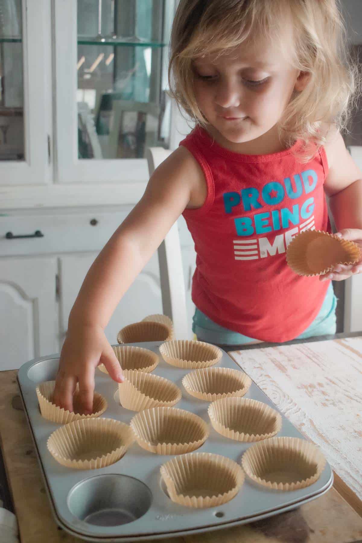 A little brunette girl helping add muffin liners to a muffin tin. 