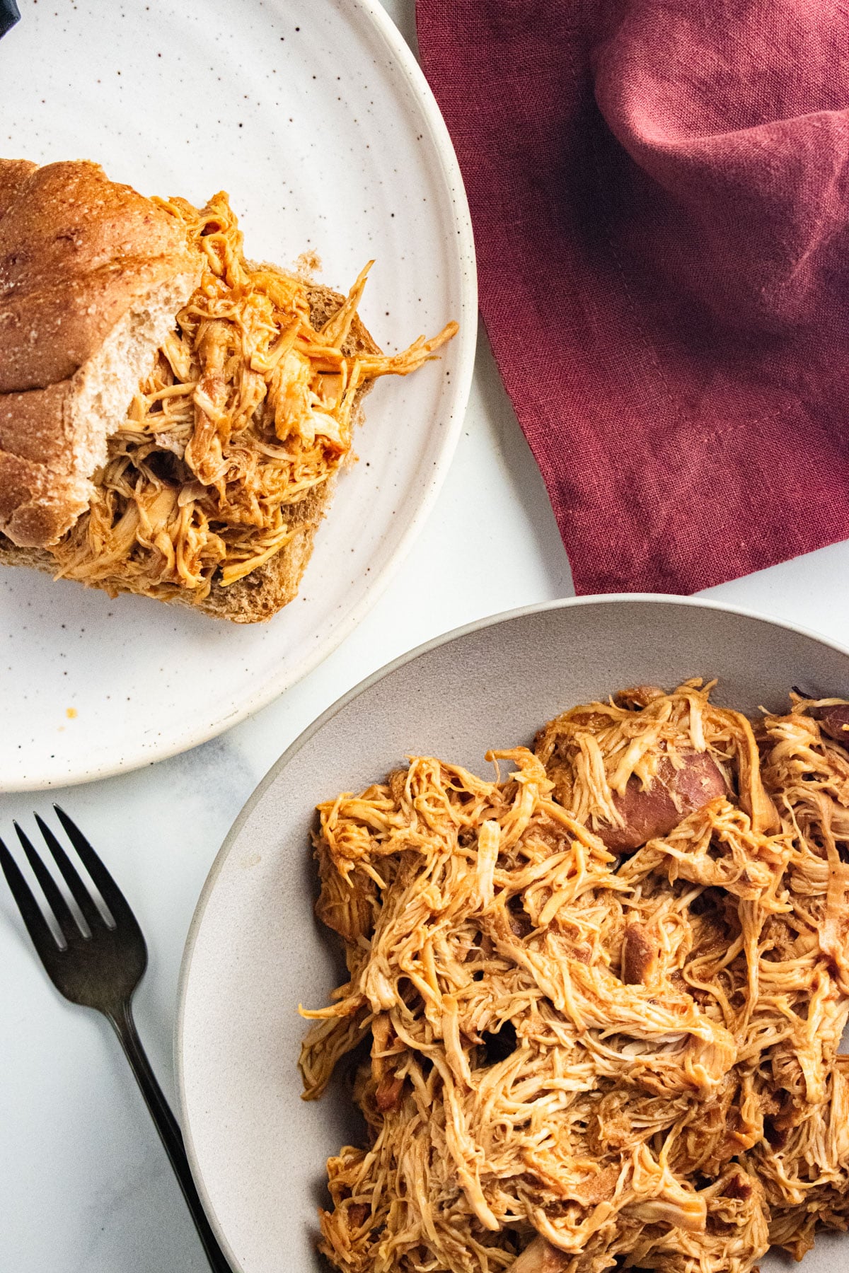Shredded BBQ chicken in a white bowl with a fork laid next to the bowl and some chicken in a bun to the side.