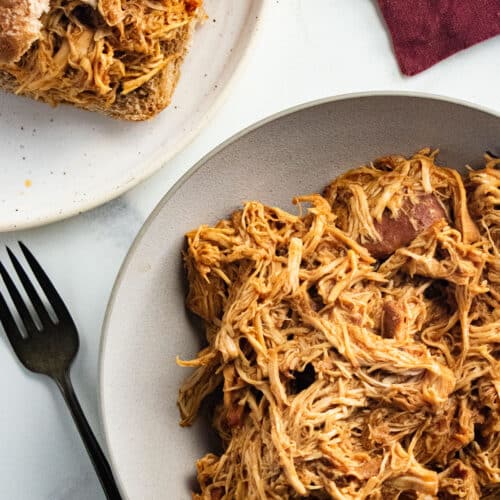 Shredded BBQ chicken in a white bowl with a fork laid next to the bowl and some of the chicken on a bun.