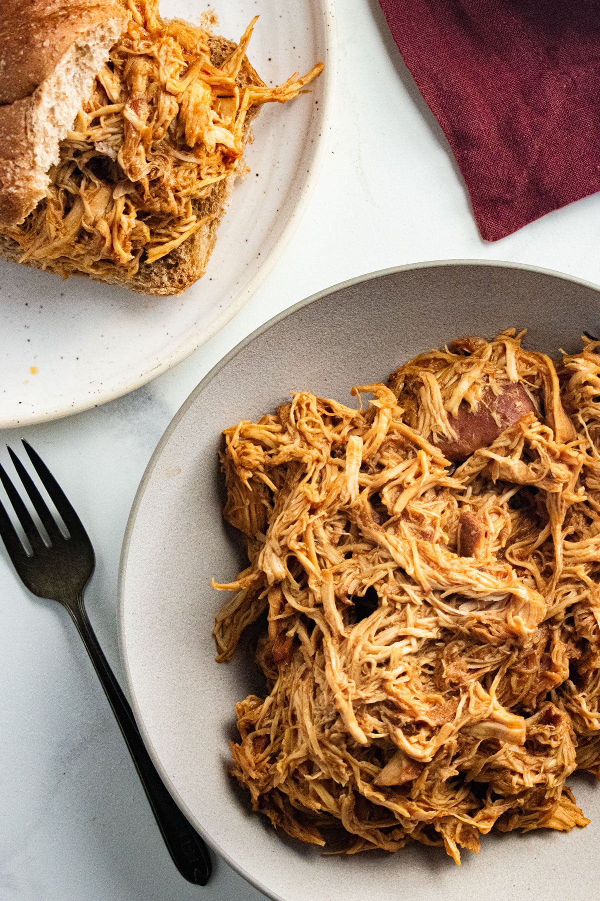 Shredded BBQ chicken in a white bowl with a fork laid next to the bowl and some of the chicken on a bun.